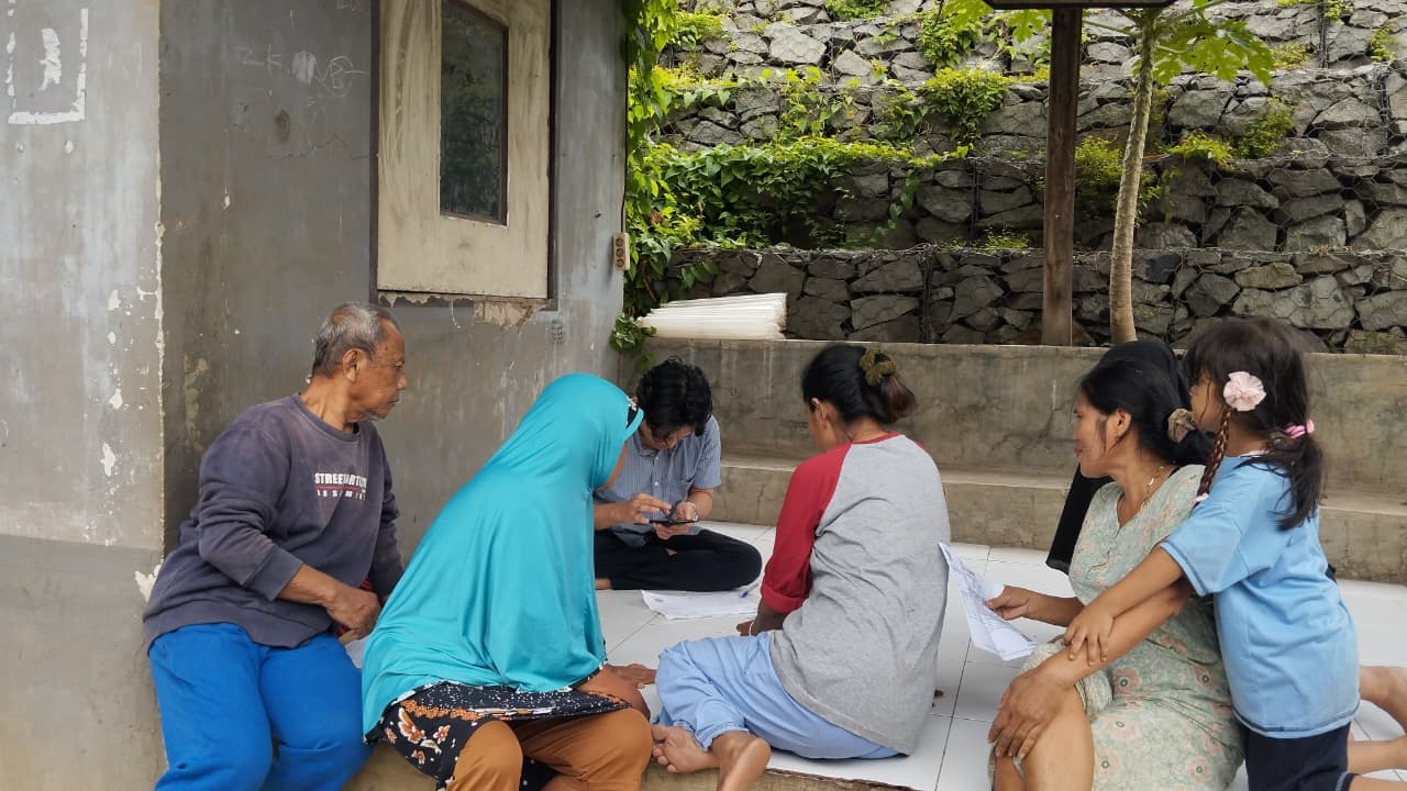 Tim lapangan duduk bersama warga di teras rumah, mencatat data sambil warga memegang formulir.