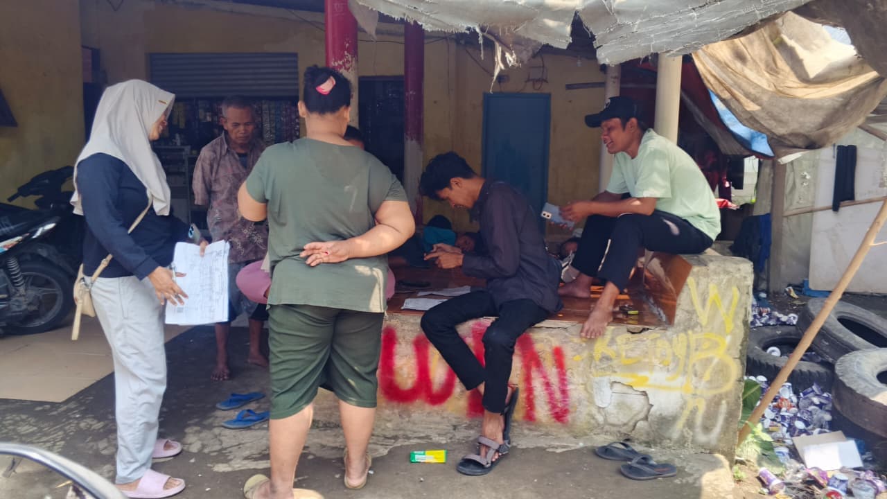 Tim lapangan memegang berkas pendataan sambil berbincang dengan warga di depan sebuah warung.