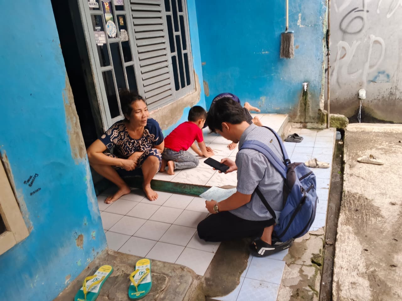 Petugas lapangan berjongkok mendata seorang ibu di teras rumahnya.